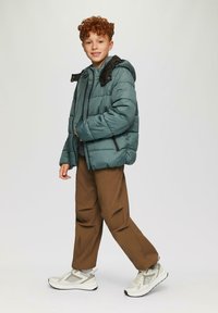 Tiefgrüne Daunenjacke mit Kapuze, schwarzen Akzenten und Reißverschlusstaschen, kombiniert mit braunen Hosen und weißen Sneakern mit Mesh- und festen Details.