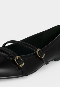 Chaussure plate noire avec deux fines brides et boucles dorées sur le dessus, texture lisse, bout arrondi, talon bas.