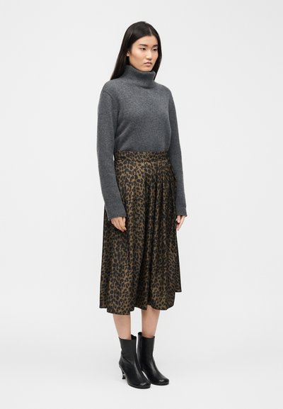 Γυναίκα που στέκεται φορώντας γκρι πουλόβερ με λαιμόκοψη, midi φούστα με σχέδιο λεοπάρ και μαύρα μποτάκια μέχρι τον αστράγαλο σε απλό φωτεινό φόντο.