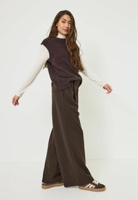Un pull sans manches en maille marron porté sur un haut à manches longues blanc, associé à un pantalon ample marron foncé et des baskets marron avec des accents blancs.