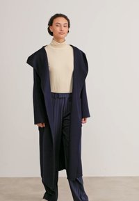 Lungo cappotto blu navy con cappuccio, materiale leggero, davanti aperto, abbinato a un dolcevita beige e pantaloni blu navy a righe.