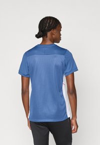 Αθλητική μπλούζα κοντού μανικιού σε μπλε χρώμα, με δίχτυ στην πλάτη, λευκά πλαϊνά και ελάχιστες ροζ λεπτομέρειες, σχεδιασμένη για άνεση και κίνηση.