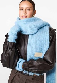 Šviesiai mėlyna raukšlėta skara, pagaminta iš švelnaus, pūkuoto medžiagos, dėvima su juoda odine striuke, turinčia raukšlėtas rankoves ir etiketę.