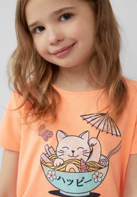 Lichtoranje t-shirt met een afbeelding van een glimlachende kat in een kom met noedels, met een parasol en bloemmotieven. Zachte stoftextuur.