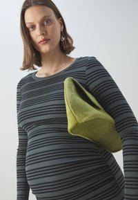 Sac en fausse fourrure verte de forme géométrique, tenu contre une personne portant un haut à manches longues ajusté noir et gris rayé.