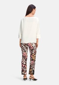Weißes, locker sitzendes Oberteil mit 3/4-Ärmeln, kombiniert mit bunten Hosen im Schmetterlingsmuster in Pink, Schwarz und Orange. Schwarze Absatzschuhe.