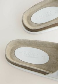 Šviesiai rudos suknelės šlepetės su baltu gumuotu padu, turinčios kontūruotą padas ir išrašytą "NEXT Forever Comfort" logotipą viduje.