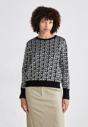 Svart strikkegenser med gjentatt "DKNY" logo mønster, kontrasterende hvit tekst, rund hals og svarte ribbede mansjetter. Brukt over et beige skjørt.