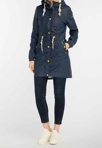 Parka bleu marine avec fermeture éclair, capuche à cordon, accents dorés et taille cintrée. Associée à un jean foncé et des baskets blanches.