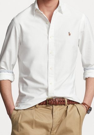 Mann trägt ein weißes Hemd mit Knöpfen und hochgekrempelten Ärmeln, beige plissierte Hose, braunen geflochtenen Gürtel und braune Lederuhr.