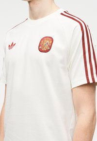 Κοντινή λήψη ενός ατόμου που φοράει λευκό αθλητικό μπλουζάκι με κόκκινες ρίγες στα μανίκια και κόκκινο-κίτρινο εθνόσημο της Ισπανίας στο στήθος.