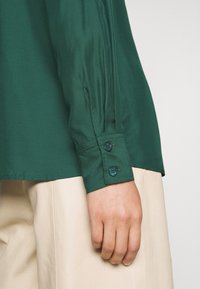 Poignet de chemise verte à manches longues avec deux boutons sur une personne portant un pantalon beige, main reposant sur le côté.