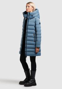 Vaaleansininen toppatakki, jossa on korkea kaulus, vetoketjusuljin ja sivutaskut. Takissa on huppu ja resorit hihansuissa. Yhdistetty mustiin leggingsseihin ja saappaisiin.