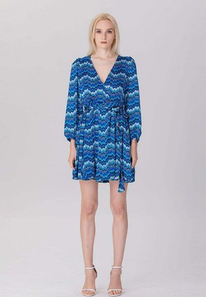 Jeune femme debout, face à l'avant, portant une robe bleu à manches longues avec un motif, un col en V et des sandales blanches à talons hauts sur un fond uni.
