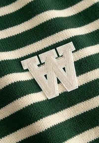 Tissu tricoté à rayures horizontales vertes et crème avec un patch blanc texturé en forme de "W" cousu dessus.