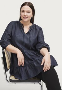Mørkeblå denim tunika med V-udskæring og samlet halsudskæring, med pufærmer og en flæset kant. Bæres over sorte leggins, mens personen sidder.