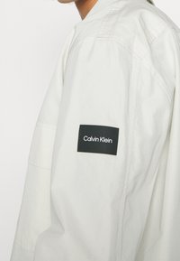 Giacca beige chiaro con una vestibilità rilassata. Presenta un'etichetta nera Calvin Klein sulla manica sinistra, realizzata in un tessuto morbido e liscio.