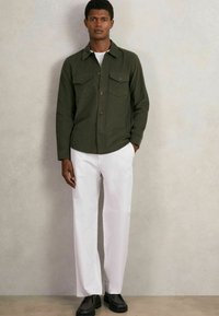 Olivgrünes Hemd mit Knopfleiste und zwei Brusttaschen, kombiniert mit weißen, geraden Hosen und schwarzen Schnürschuhen. Glatte Textur und moderner Schnitt.
