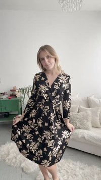 Moteris juodo gėlėto suknele, laikanti sijoną, šviesiame svetainės kambaryje, už jos matyti balta sofa ir žalia spintelė.