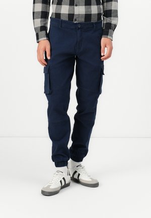 Homme portant un pantalon cargo jogger bleu marine, des chaussettes blanches, des baskets blanches avec des rayures noires, et une chemise à carreaux noir et gris boutonnée.