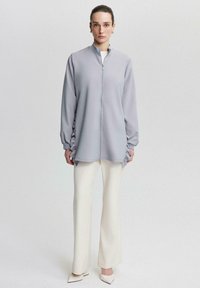 Jacheta gri cu fermoar, cu mâneci lungi și laterale strânse, asociată cu pantaloni evazați crema și pantofi cu vârful ascuțit. Textură de țesătură netedă.