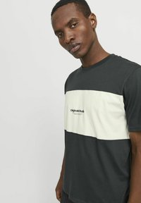 Camiseta de manga corta con rayas horizontales en gris oscuro y beige claro, que presenta el logo "Originals Studio" en negro en el centro.