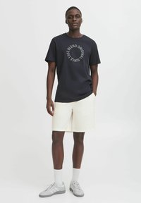 Marineblå t-shirt med cirkulært hvidt grafisk tekst, parret med beige shorts, hvide sokker og grå sneakers. Stærkt bomuldsstof, afslappet pasform.