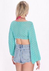 Brassière en crochet de couleur turquoise clair avec de longues manches amples et des motifs géométriques, associée à un short en denim aux bords effilochés.