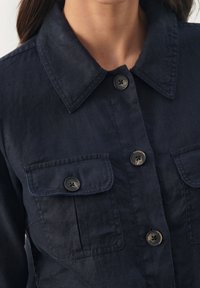 Camisa de lino azul marino oscuro con botones en la parte delantera, cuello puntiagudo y dos bolsillos en el pecho. Presenta botones negros y un acabado de tejido texturizado.