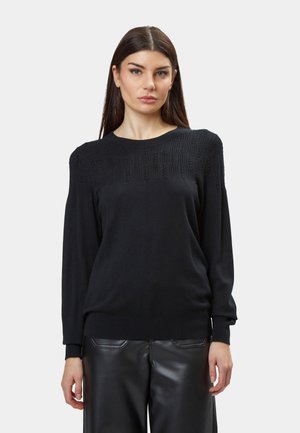 Schwarzer Strickpullover mit einem strukturierten Muster an den Schultern, langen Ärmeln und einem gerippten Saum. Tragen mit hochgeschnittenen schwarzen Lederhosen.