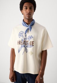 Cremefarvet t-shirt med korte ærmer, der har blå blomstergraphics og teksten "Breathe" i brun. Bæres med et blåt mønstret tørklæde.