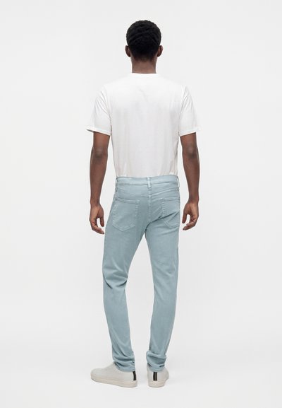 Homme debout de dos, portant un t-shirt blanc uni, un jean bleu clair et des baskets blanches sur un fond clair uni.