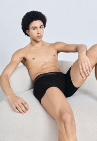 Sorte boksertrusser med en TOMMY HILFIGER logo på linningen, båret af en person, der sidder på en lys, tekstureret, rund sofa mod en ensfarvet baggrund.
