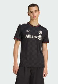 Young man wearing black Adidas sports jersey with Allianz logo and red accents, standing against plain light gray background.