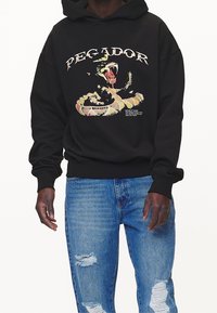 Černá mikina s kapucí s velkou grafikou hada a nápisy na přední straně. Nošeno s světle modrými džíny s vyšisovaným vzhledem. Měkká látka, volný střih.