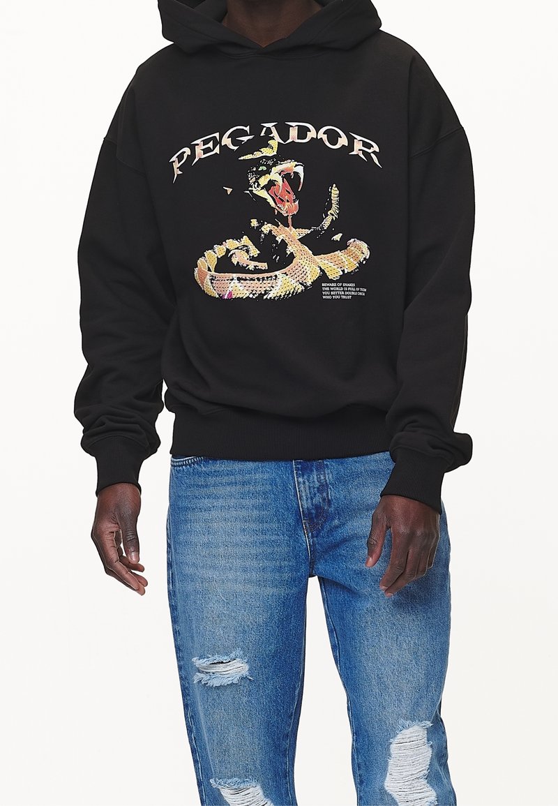 Černá mikina s kapucí s velkou grafikou hada a nápisy na přední straně. Nošeno s světle modrými džíny s vyšisovaným vzhledem. Měkká látka, volný střih.