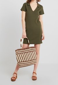 Olivgrünes geripptes Kleid mit kurzen Ärmeln und V-Ausschnitt. Geflochtete Tasche in Braun und Creme mit geschwungenen Griffen. Braune Plateausandaletten.