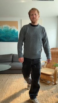 Suéter cinza tricotado com gola redonda, combinado com jeans escuros e ténis cinzas. Ambiente interior com uma cadeira trançada e obras de arte.