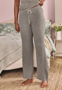 Šedé žebrované volnočasové kalhoty s pružným pasem na stažení, širokými nohavicemi a měkkou texturou. Model stojí bosý na tkaném koberci.