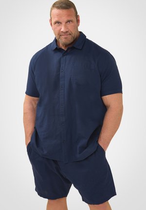 Mand med skæg står med hænderne i lommerne iført en marineblå kortærmet knap-op skjorte og matchende shorts mod en hvid baggrund.