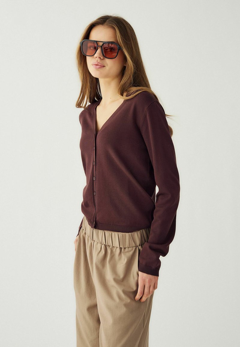 Mladá žena s velkými čtvercovými slunečními brýlemi, hnědým propínacím svetrem a béžovými volnými kalhotami na jednoduchém světlém pozadí.