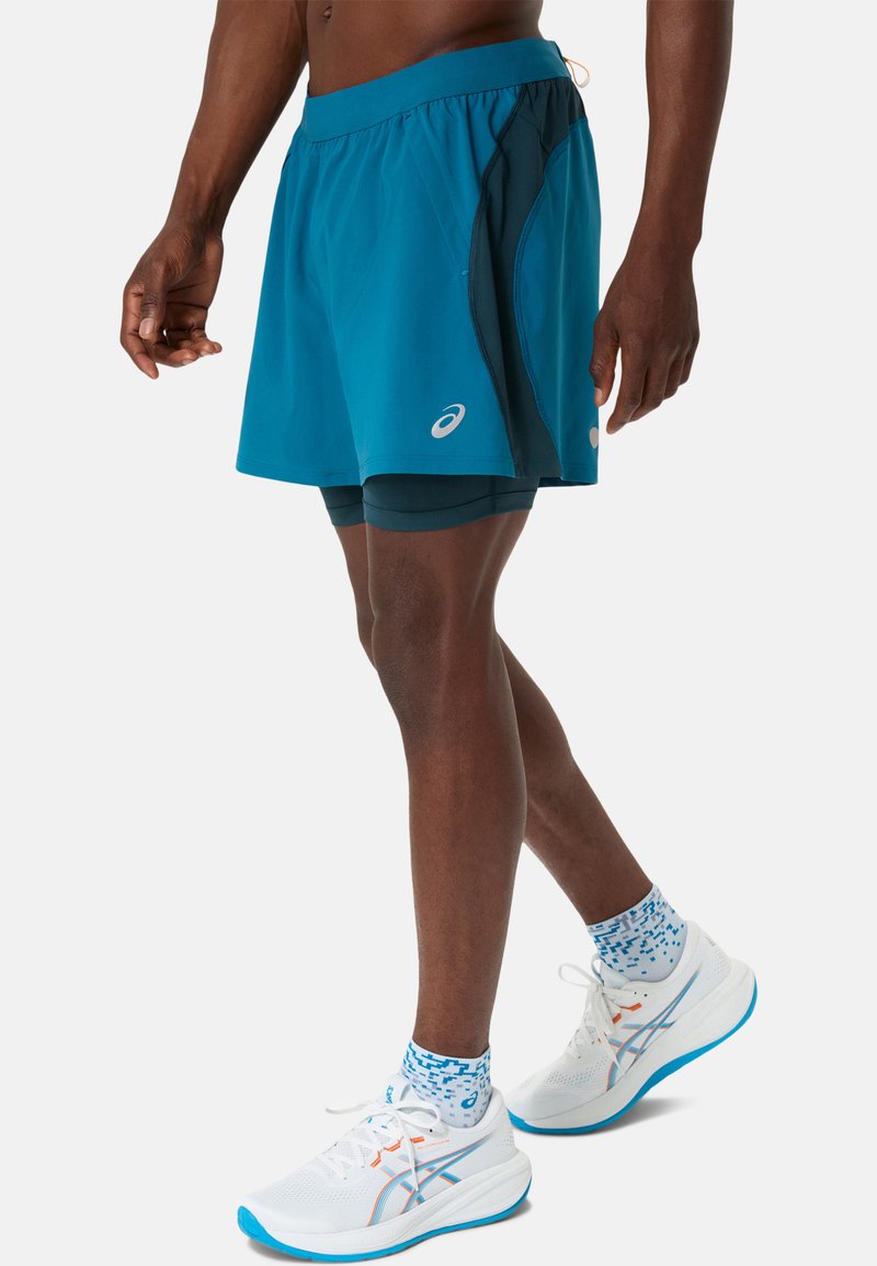 Shorts de sport teal avec des accents noirs, taille élastique et doublure intérieure. Associés à des chaussures de course blanches et des chaussettes bleues à motifs.