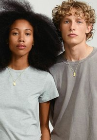 Två personer står nära varandra, bärande t-shirts med kort ärm: en grå och en ljusgrå. Båda har på sig guldhalsband med berlocker.