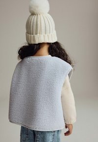 Lichtblauw fleece vest met ronde schouders, gedragen over een crèmekleurig shirt met lange mouwen. Witte gebreide muts met een fluffy pompon bovenop.