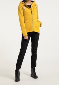Gelbe Fleece-Zip-Jacke mit Kapuze, kontrastierender marineblauer Fütterung, zwei Fronttaschen und Kordelzug-Details. Dazu eine schwarze, eng anliegende Hose und Stiefel.