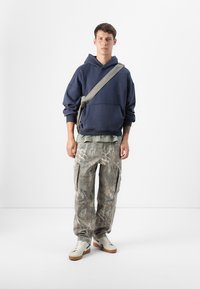 Hanorac oversized bleumarin cu un strat inferior verde, pantaloni cargo cu imprimeu de camuflaj și adidași albi. Include o geantă crossbody și materiale texturate.