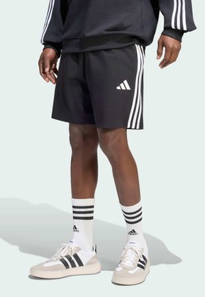 Osoba nosząca czarne szorty Adidas z białymi paskami, białe skarpetki Adidas z czarnymi paskami oraz beżowe buty sportowe Adidas z czarnymi paskami.