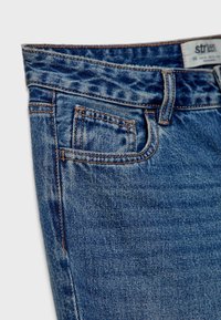 Gros plan sur un jean en denim bleu montrant la poche avant, la boucle de ceinture et les détails de couture visibles.