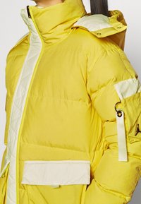 Keltainen untuvatakki, jossa korkea kaulus, kermaiset yksityiskohdat, etutasku ja sivutapin metallikoriste. Sileä pinta.