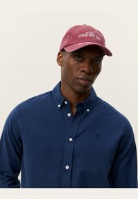 Maroon cap with "Lower West Side" embroidery, cotton material. Worn with a blue button-up shirt, showcasing a textured fabric.